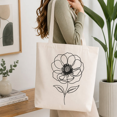 A woman wearing a sage green ribbed sweater carries a cream canvas tote bag with a black line art flower design. The scene is set in a bright, modern room with a wooden table, books, a small vase, and a large potted plant.