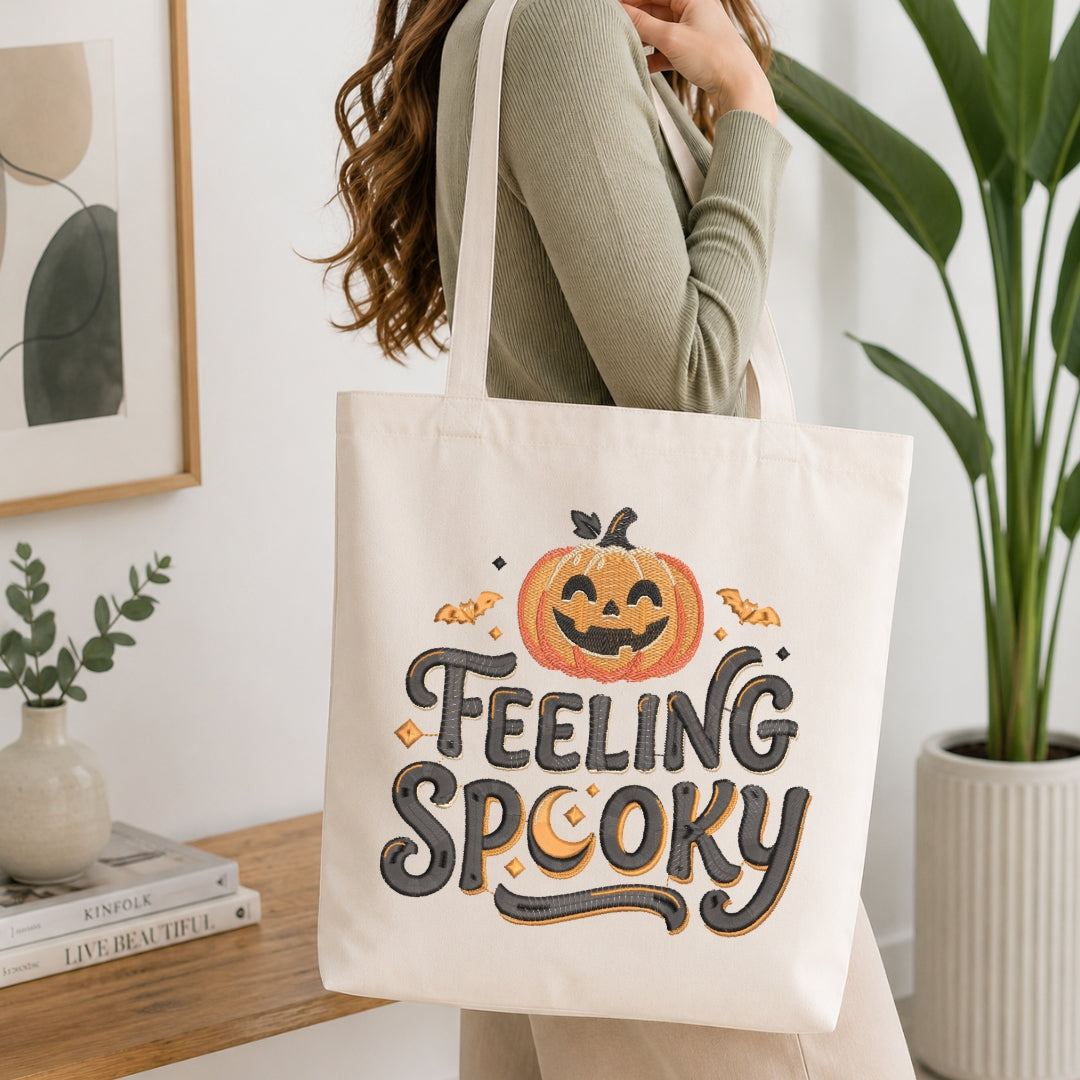 A woman wearing a ribbed sage green long-sleeve top carries a cream canvas tote bag with a 'FEELING SPOOKY' Halloween graphic. The tote bag features an orange jack-o'-lantern, bats, and a crescent moon with black and gray lettering.