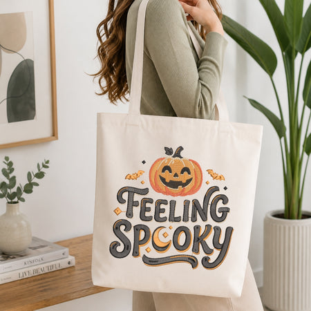 A woman wearing a ribbed sage green long-sleeve top carries a cream canvas tote bag with a 'FEELING SPOOKY' Halloween graphic. The tote bag features an orange jack-o'-lantern, bats, and a crescent moon with black and gray lettering.