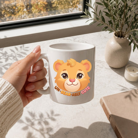A hand holding a white ceramic coffee mug featuring a cute cartoon bear face illustration and text Un-Bear-Ably Cute on a sunny marble surface.
