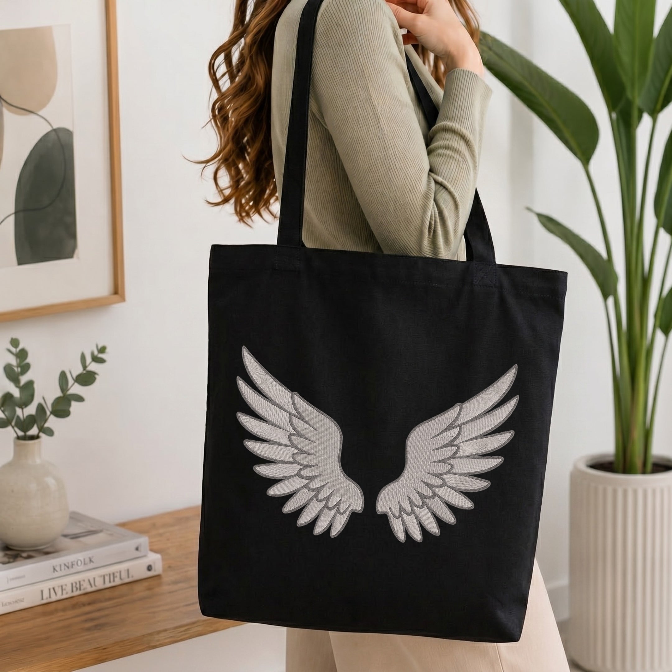 A woman wearing a beige ribbed sweater carries a black canvas tote bag with a large white embroidered angel wings design on the front, set in a bright, modern room with a wooden table, books, and a potted plant.