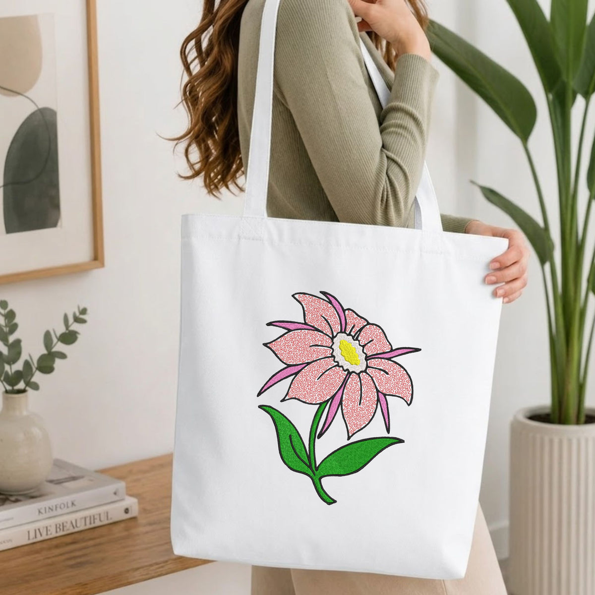 Woman carrying a white natural canvas tote bag featuring an embroidered pink flower design in a bright, modern interior setting