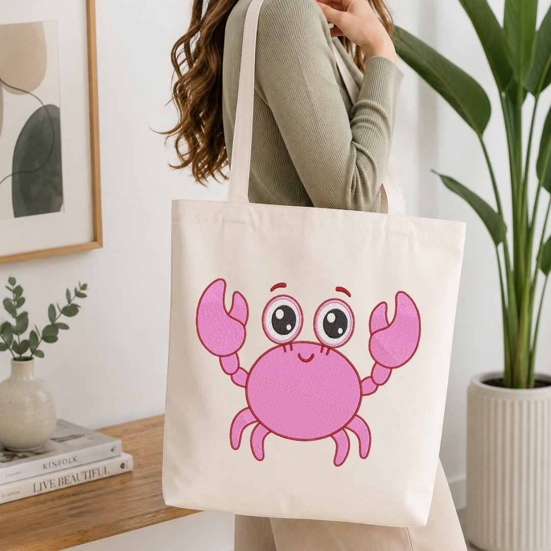 A woman wearing a sage green ribbed sweater carries a cream canvas tote bag featuring a cute pink cartoon crab graphic. The scene is set in a bright, modern room with a wooden table, books, a small vase, and a potted plant.