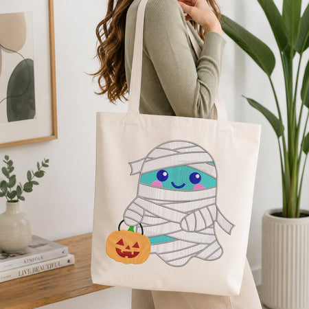 A person wearing a sage green ribbed long-sleeve top carries a cream canvas tote bag. The bag features a cute cartoon graphic of a smiling ghost mummy wrapped in white bandages, holding an orange jack-o'-lantern pumpkin.