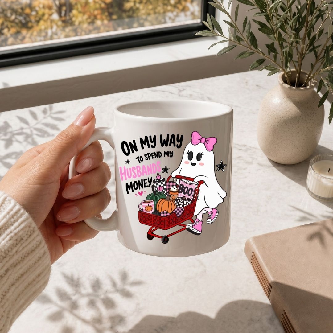 A person holding a white ceramic coffee mug printed with the "On My Way to Spend My Husbands Money" ghost Halloween illustration on a white marble table.