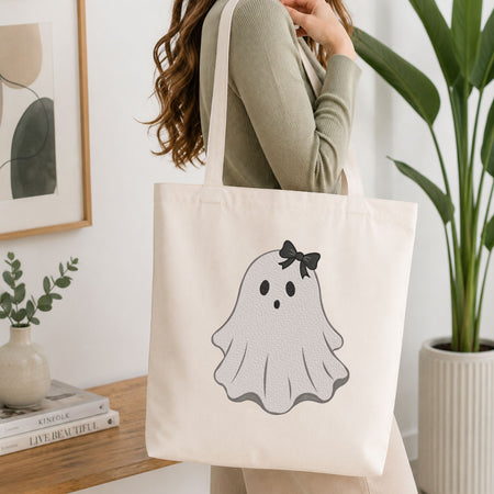 Woman carrying a white natural canvas tote bag featuring a cute ghost machine embroidery design in a bright, modern interior setting   