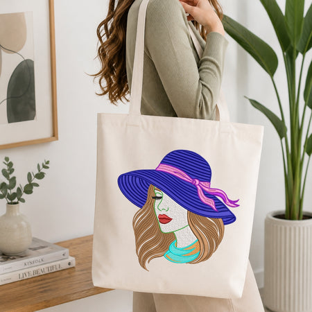 A woman in a ribbed sage green top carries a cream canvas tote bag with a colorful graphic of a woman wearing a purple sunhat with a pink ribbon. The scene is set in a bright, modern room with a wooden table, books, and a potted plant.