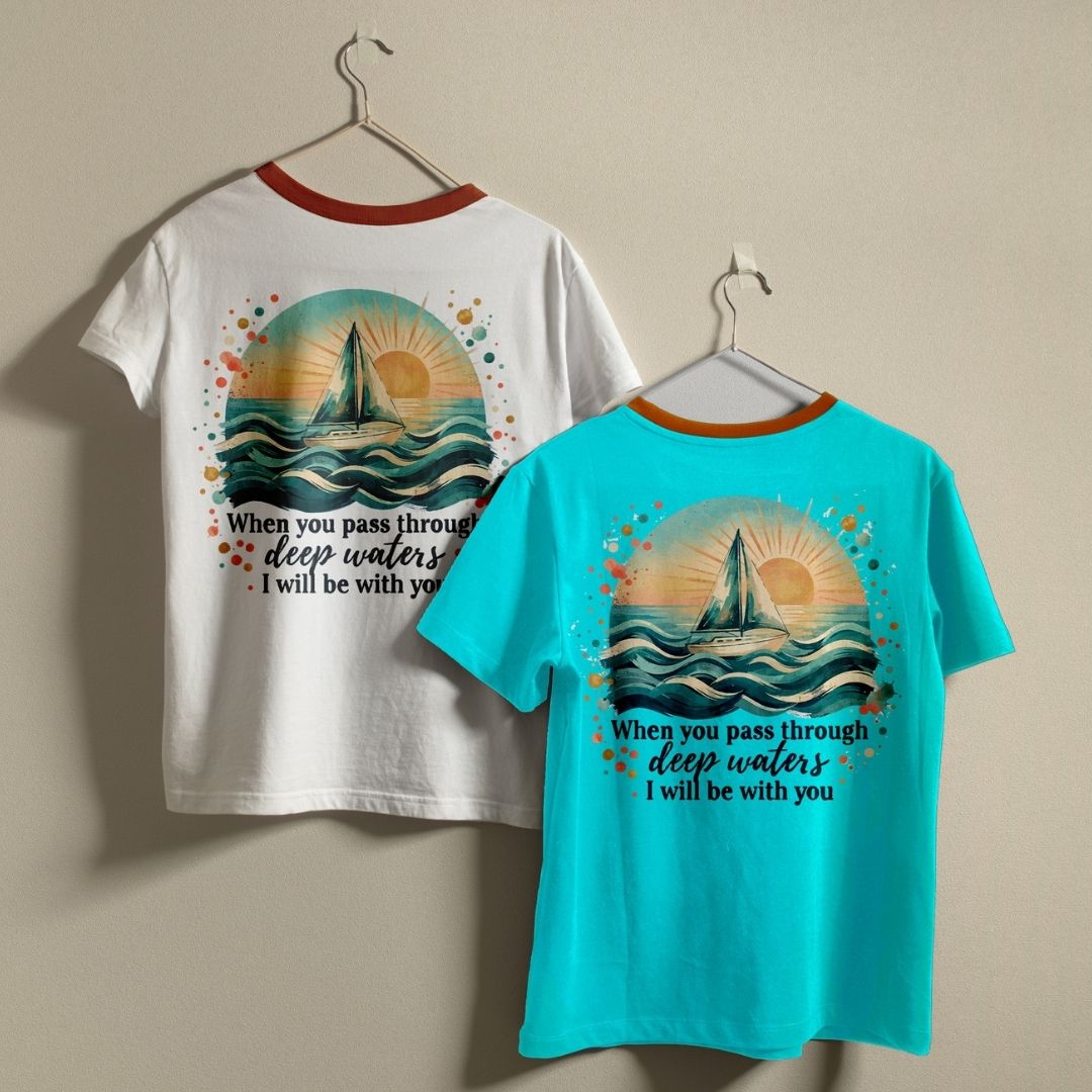 Two ringer t-shirts hanging on hangers against a beige wall. One shirt is white with a red collar and shows the back graphic, which features a sailboat on waves and the text 'When you pass through deep waters I will be with you'.