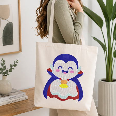 A woman holds a cream canvas tote bag featuring a colorful embroidered cartoon penguin character. The setting is a bright, modern room with a wooden table, books, a framed print, and a potted plant in the background.