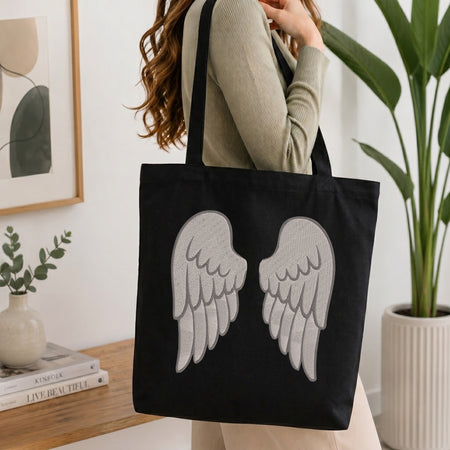A woman wearing a beige ribbed sweater carries a black canvas tote bag with a large gray embroidered angel wings design on the front, set in a bright, modern room with a wooden table, framed art, and a potted plant.