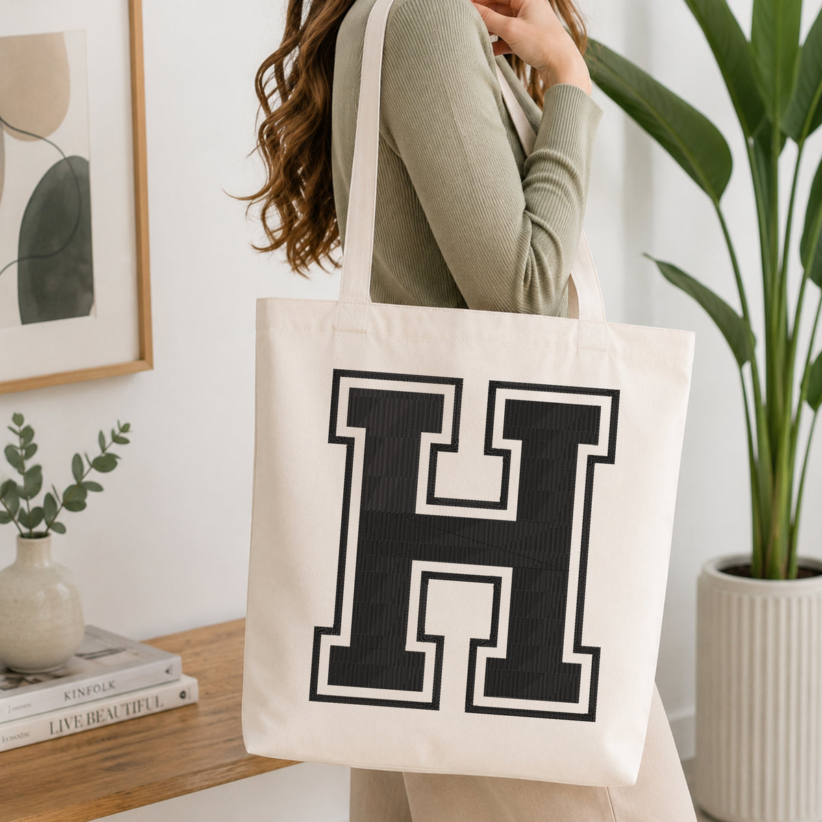 A woman wearing a sage green ribbed long-sleeve top carries a large off-white canvas tote bag with a bold black block letter 'H' outlined in white, shown in a bright, modern room with a wooden console table and a potted plant.
