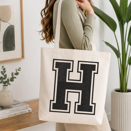 A woman wearing a sage green ribbed long-sleeve top carries a large off-white canvas tote bag with a bold black block letter 'H' outlined in white, shown in a bright, modern room with a wooden console table and a potted plant.