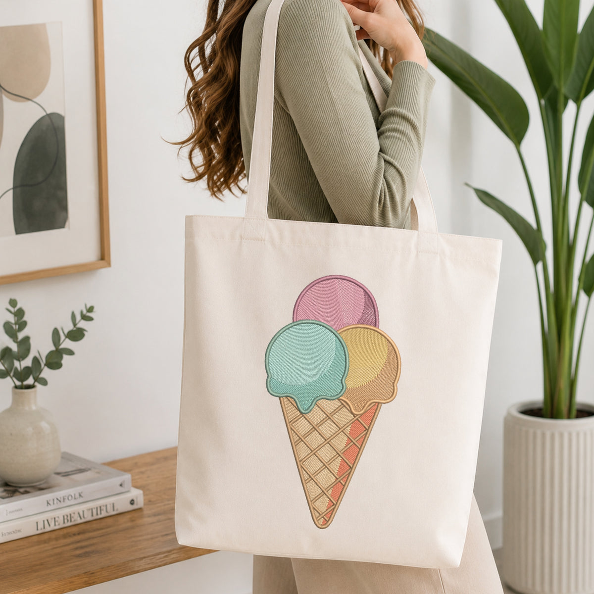A woman wearing a sage green ribbed long-sleeve top carries a cream canvas tote bag with a colorful embroidered ice cream cone design. The setting is a bright, modern room with a wooden table, framed art, and a potted plant.