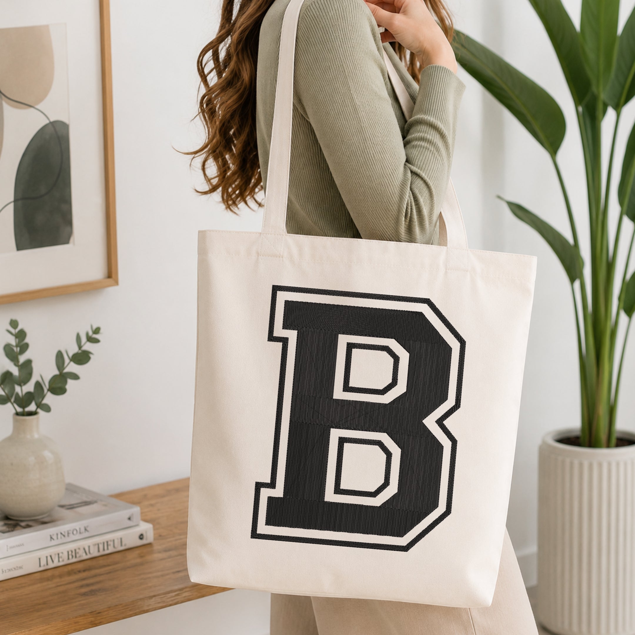 A woman wearing a sage green ribbed long-sleeve top carries a large off-white canvas tote bag with a bold black collegiate letter 'B' outlined in white. The scene is set in a bright, modern room with a wooden console table, a framed print, a small vase with greenery, and a potted plant.