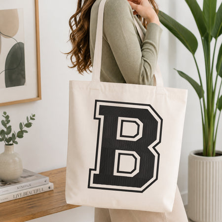 A woman wearing a sage green ribbed long-sleeve top carries a large off-white canvas tote bag with a bold black collegiate letter 'B' outlined in white. The scene is set in a bright, modern room with a wooden console table, a framed print, a small vase with greenery, and a potted plant.