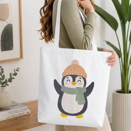 A woman wearing a sage green ribbed sweater carries a white canvas tote bag featuring a cute cartoon penguin wearing a tan knit hat and a green scarf. The scene is set in a bright, modern room with a wooden table, books, and a potted plant.