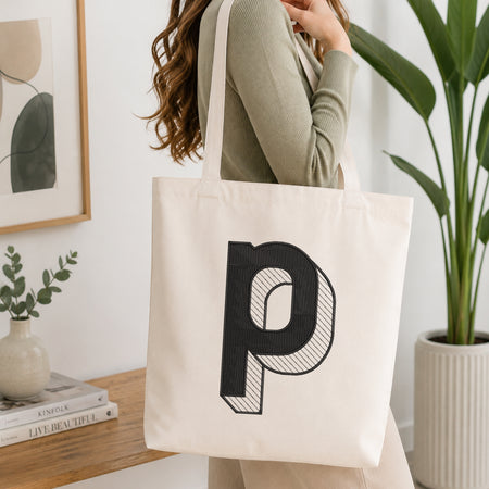 A woman wearing a sage green ribbed long-sleeve top carries a large off-white canvas tote bag with a bold black letter 'p' printed on the front, set in a bright, modern room with a wooden table, framed art, and a potted plant.