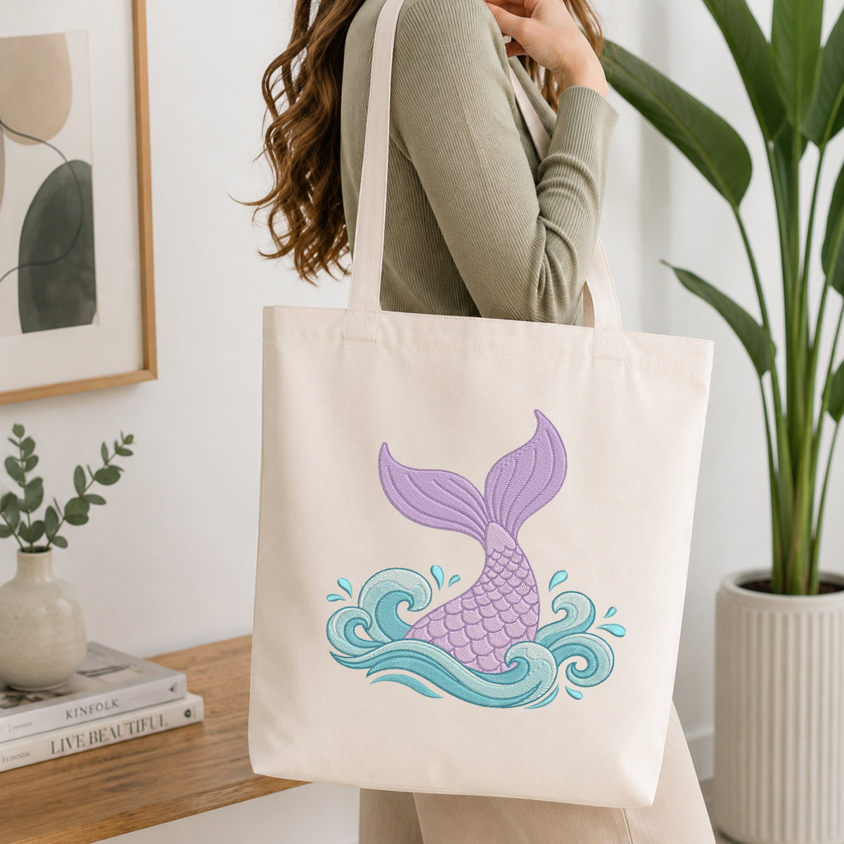 Woman wearing a green top holding a natural canvas tote bag featuring a stitched purple and blue mermaid tail design in a bright room.