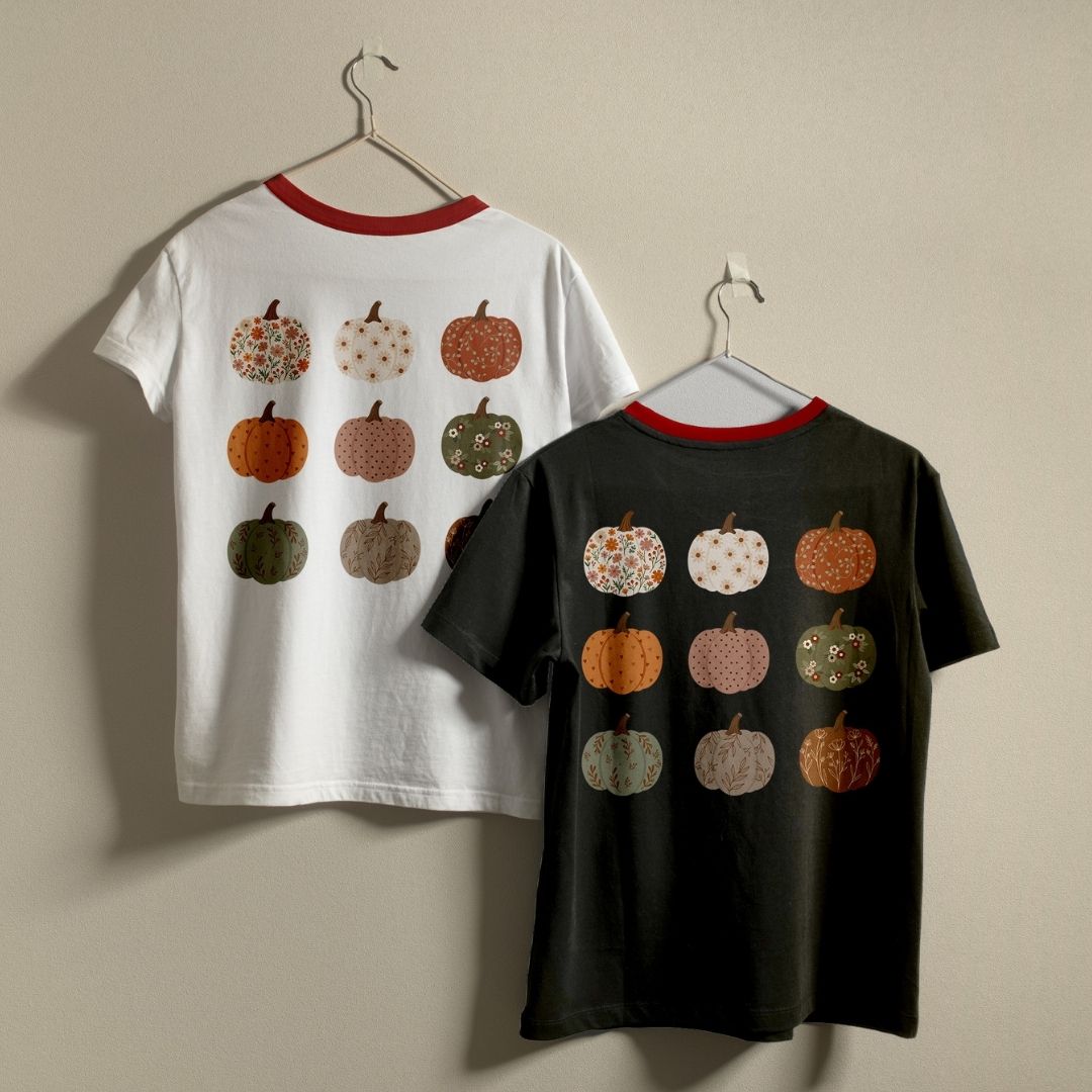 Two short-sleeve ringer T-shirts hanging on hangers against a plain beige wall. The left shirt is white with a red collar and features a back print of nine patterned pumpkins in orange, green, brown, and cream.