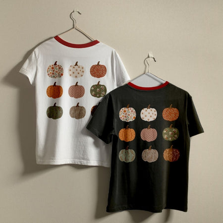 Two short-sleeve ringer T-shirts hanging on hangers against a plain beige wall. The left shirt is white with a red collar and features a back print of nine patterned pumpkins in orange, green, brown, and cream.