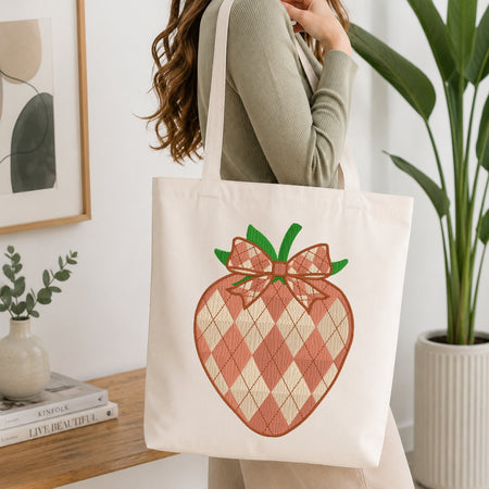 An off-white canvas tote bag with a large embroidered strawberry design in pink and beige argyle with green leaves, carried by a woman in a ribbed sage green top in a bright, modern room.