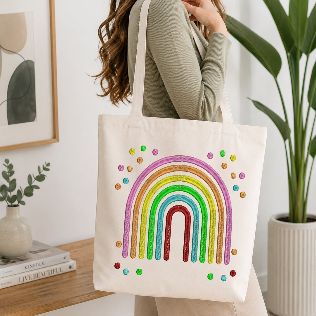A woman wearing a sage green ribbed sweater carries a cream canvas tote bag decorated with a colorful glitter rainbow and small rainbow dots. The scene is set in a bright, modern room with a wooden table, books, a potted plant, and framed wall art.