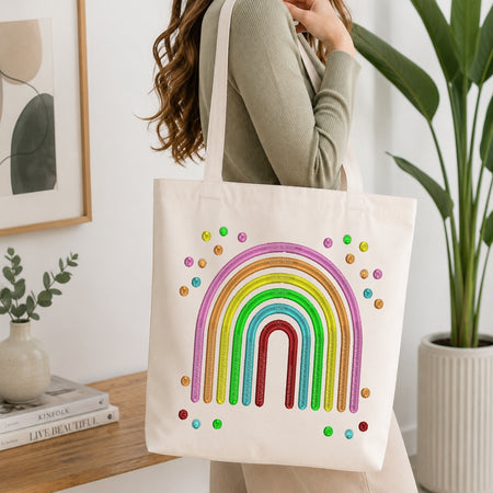 A woman wearing a sage green ribbed sweater carries a cream canvas tote bag decorated with a colorful glitter rainbow and small rainbow dots. The scene is set in a bright, modern room with a wooden table, books, a potted plant, and framed wall art.