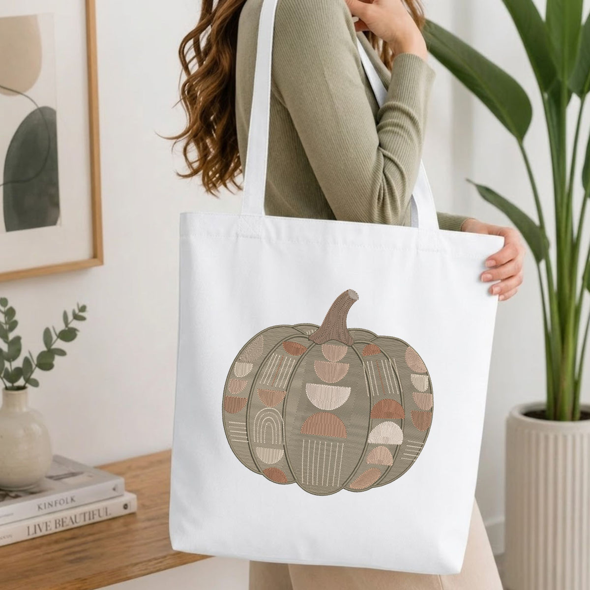  Woman carrying a white natural canvas tote bag featuring an embroidered geometric pumpkin design in a bright, modern interior setting.   