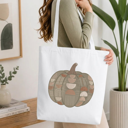  Woman carrying a white natural canvas tote bag featuring an embroidered geometric pumpkin design in a bright, modern interior setting.   