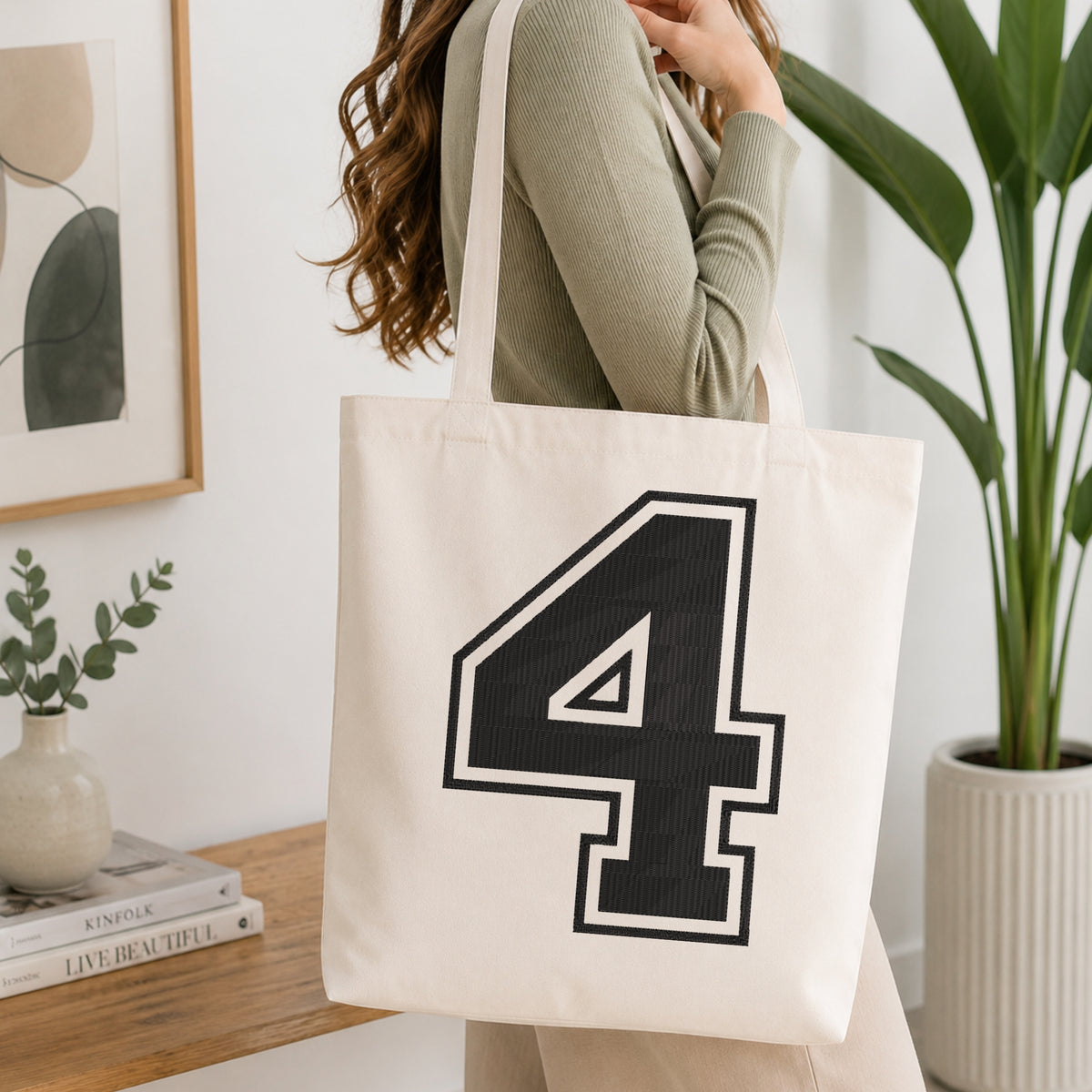 A woman wearing a sage green ribbed long-sleeve top carries a large off-white canvas tote bag with a bold black number 4 outlined in white on the front, set in a bright, modern room with a wooden console table, framed art, and a potted plant.