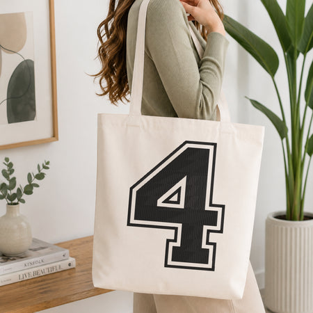 A woman wearing a sage green ribbed long-sleeve top carries a large off-white canvas tote bag with a bold black number 4 outlined in white on the front, set in a bright, modern room with a wooden console table, framed art, and a potted plant.