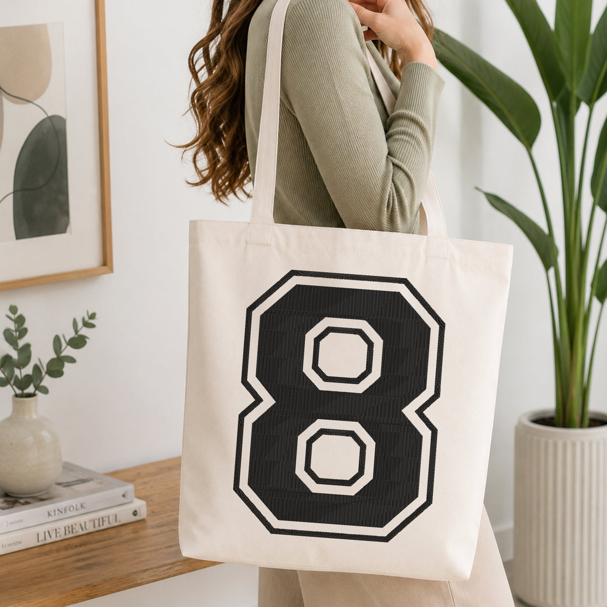 A woman wearing a sage green ribbed long-sleeve top carries a cream canvas tote bag with a large black and white number 8 graphic on the front, set in a bright, modern room with a wooden console table and a potted plant.