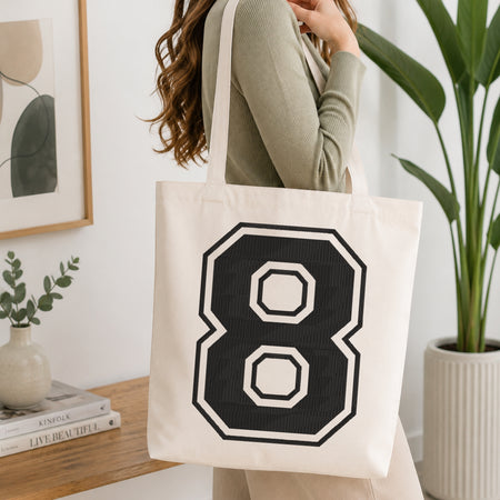 A woman wearing a sage green ribbed long-sleeve top carries a cream canvas tote bag with a large black and white number 8 graphic on the front, set in a bright, modern room with a wooden console table and a potted plant.