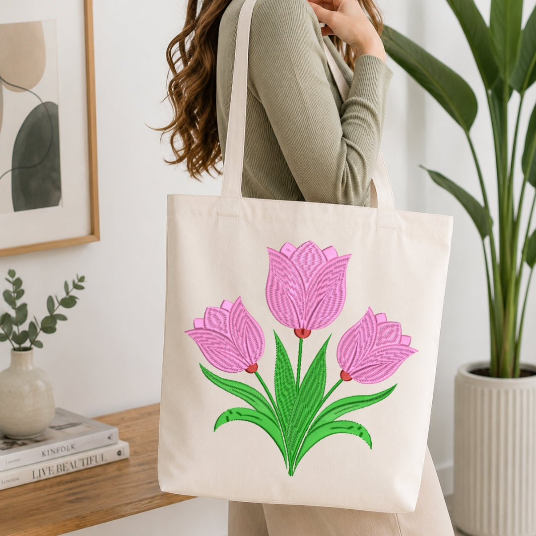 A person wearing a ribbed sage green long-sleeve top carries a cream canvas tote bag with a large embroidered design of three pink tulips with green leaves. The scene is set in a bright, modern room with a wooden table, books, a small vase, and a potted plant.
