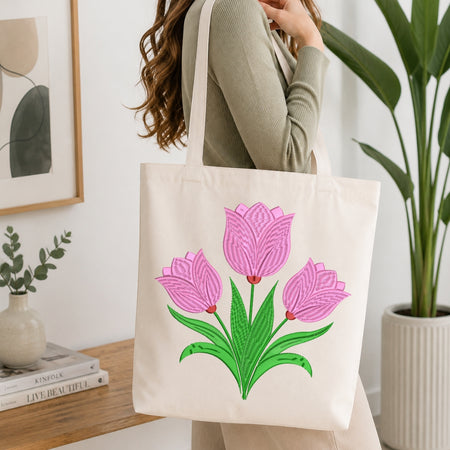 A person wearing a ribbed sage green long-sleeve top carries a cream canvas tote bag with a large embroidered design of three pink tulips with green leaves. The scene is set in a bright, modern room with a wooden table, books, a small vase, and a potted plant.