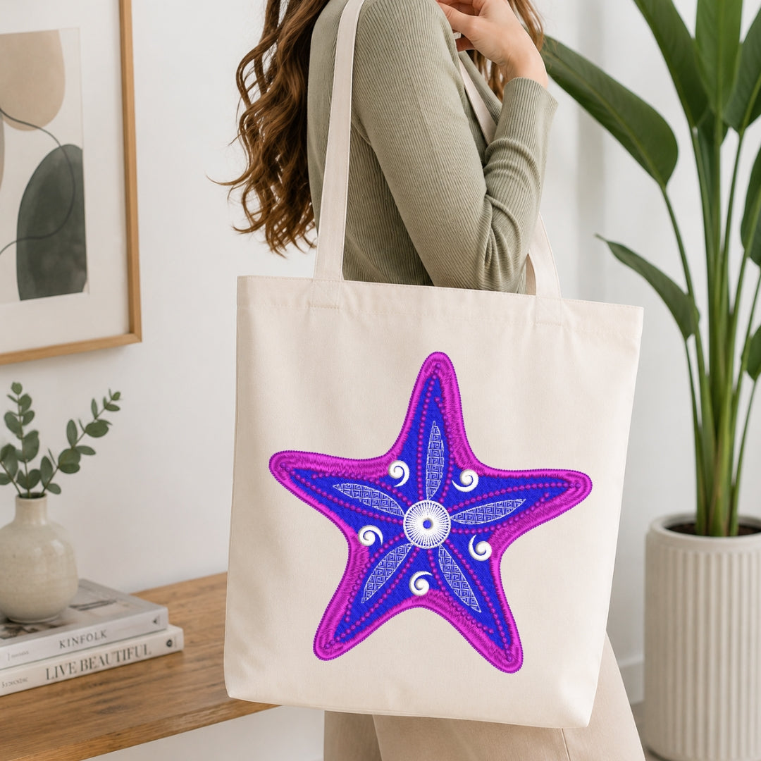 A woman wearing a sage green ribbed long-sleeve top carries a cream canvas tote bag with a large embroidered starfish design in purple and blue on the front, set in a bright, modern room with a wooden table and potted plants.