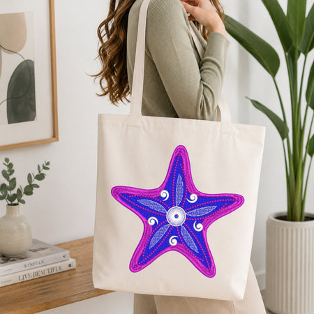 A woman wearing a sage green ribbed long-sleeve top carries a cream canvas tote bag with a large embroidered starfish design in purple and blue on the front, set in a bright, modern room with a wooden table and potted plants.