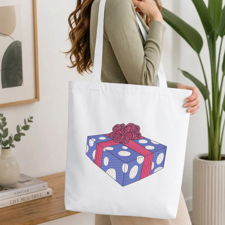 Woman carrying a white natural canvas tote bag featuring an embroidered wrapped gift box design in a bright, modern interior setting