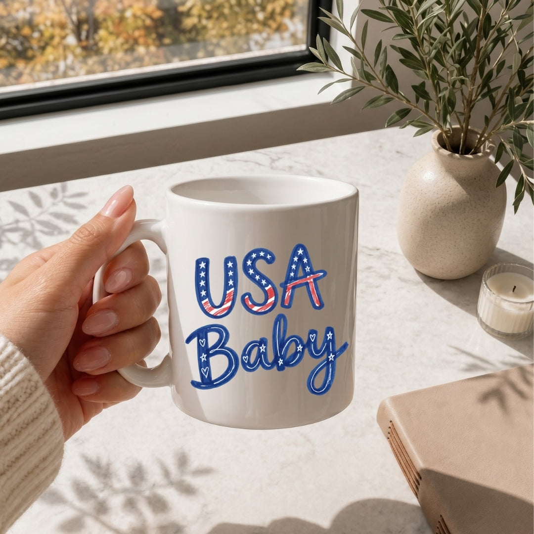 Person holding a mug with 'USA Baby' text on a table with a plant and notebook.
