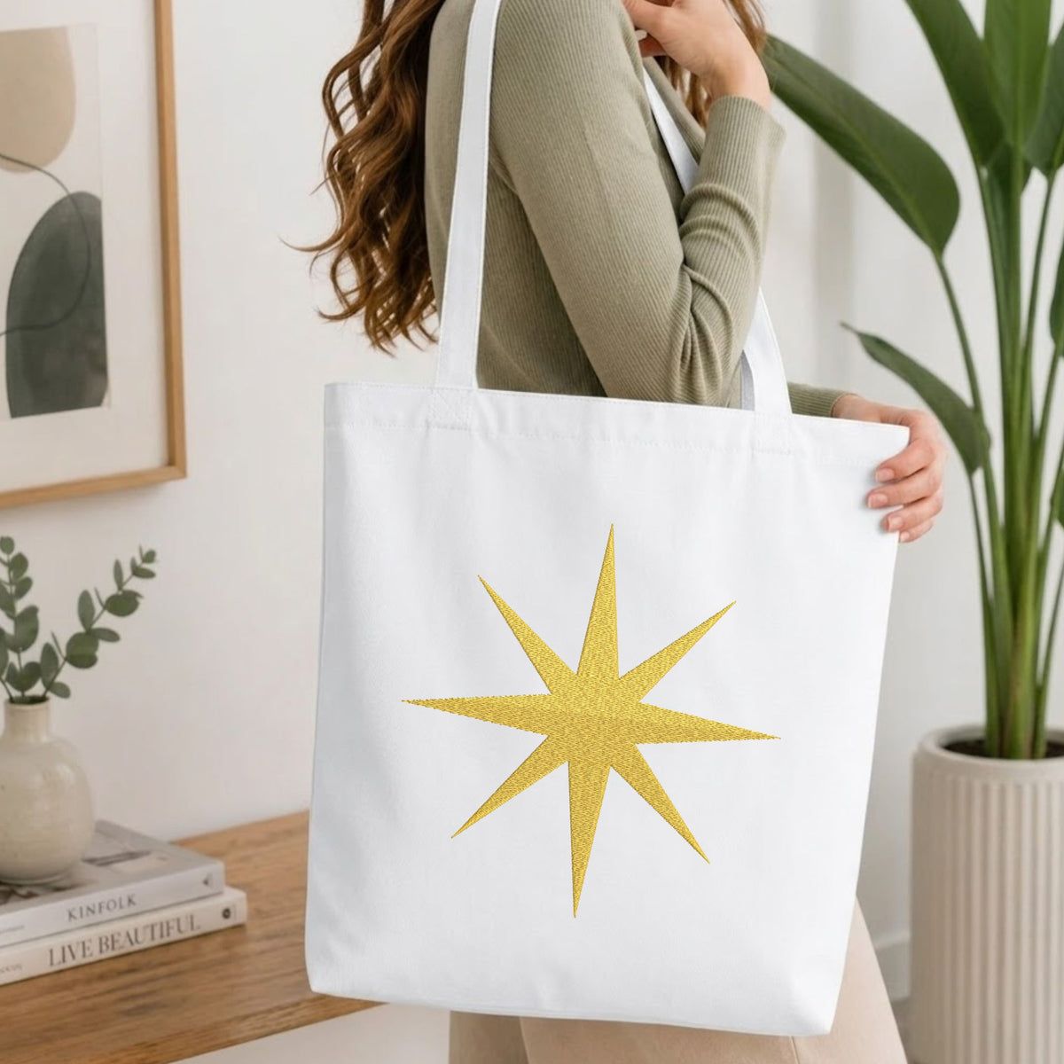 A woman wearing a sage green ribbed long-sleeve top carries a white canvas tote bag with a glittery gold 12-point star design on the front, set in a bright, modern room with a wooden table, books, and a potted plant.