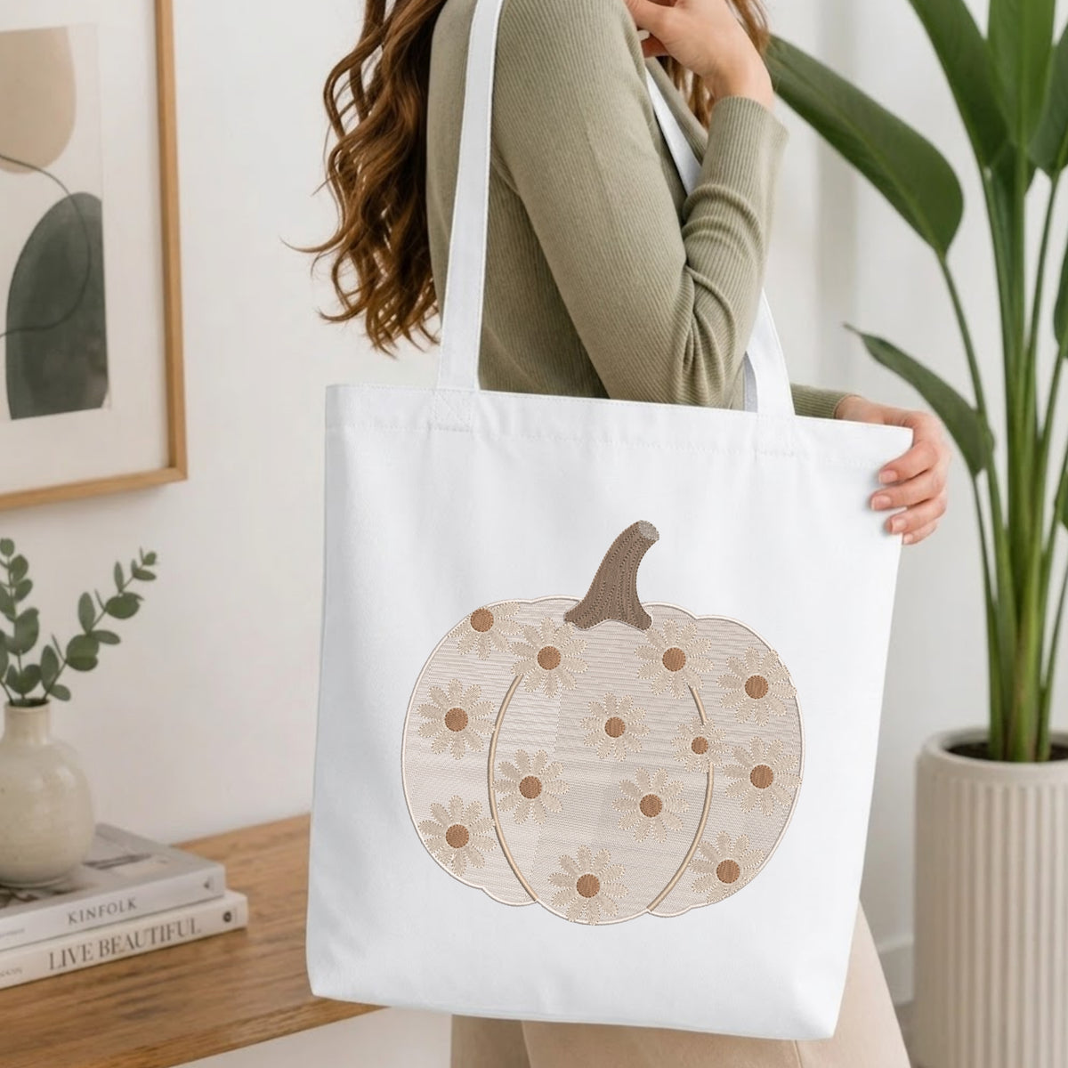 A woman wearing a sage green ribbed long-sleeve top carries a white canvas tote bag with a large pumpkin graphic. The pumpkin design features a brown stem, beige and white daisy patterns, and small brown dots.