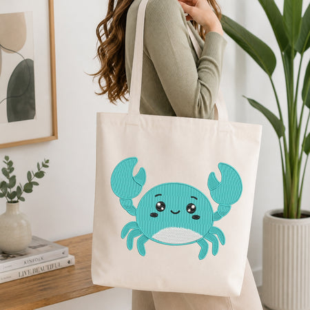 An off-white canvas tote bag with a cute embroidered teal and white crab design, carried by a woman wearing a sage green ribbed sweater in a bright, modern room.