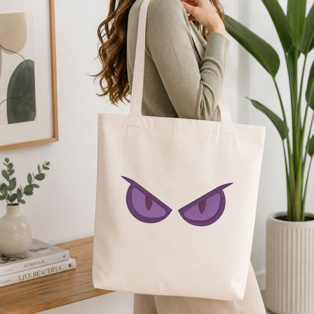 A woman wearing a sage green ribbed top carries a cream canvas tote bag featuring a graphic of two angry purple eyes. The setting is a bright, modern room with a wooden table, books, a framed print, and a potted plant.