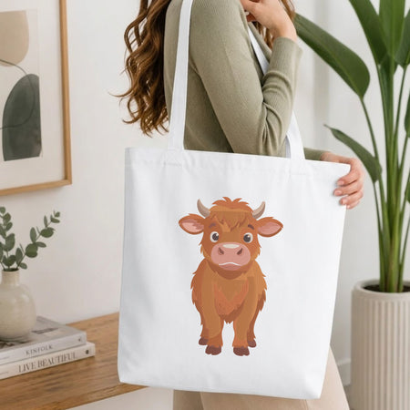 Woman carrying a white natural canvas tote bag featuring a cute Highland cow machine embroidery design in a bright, modern interior setting