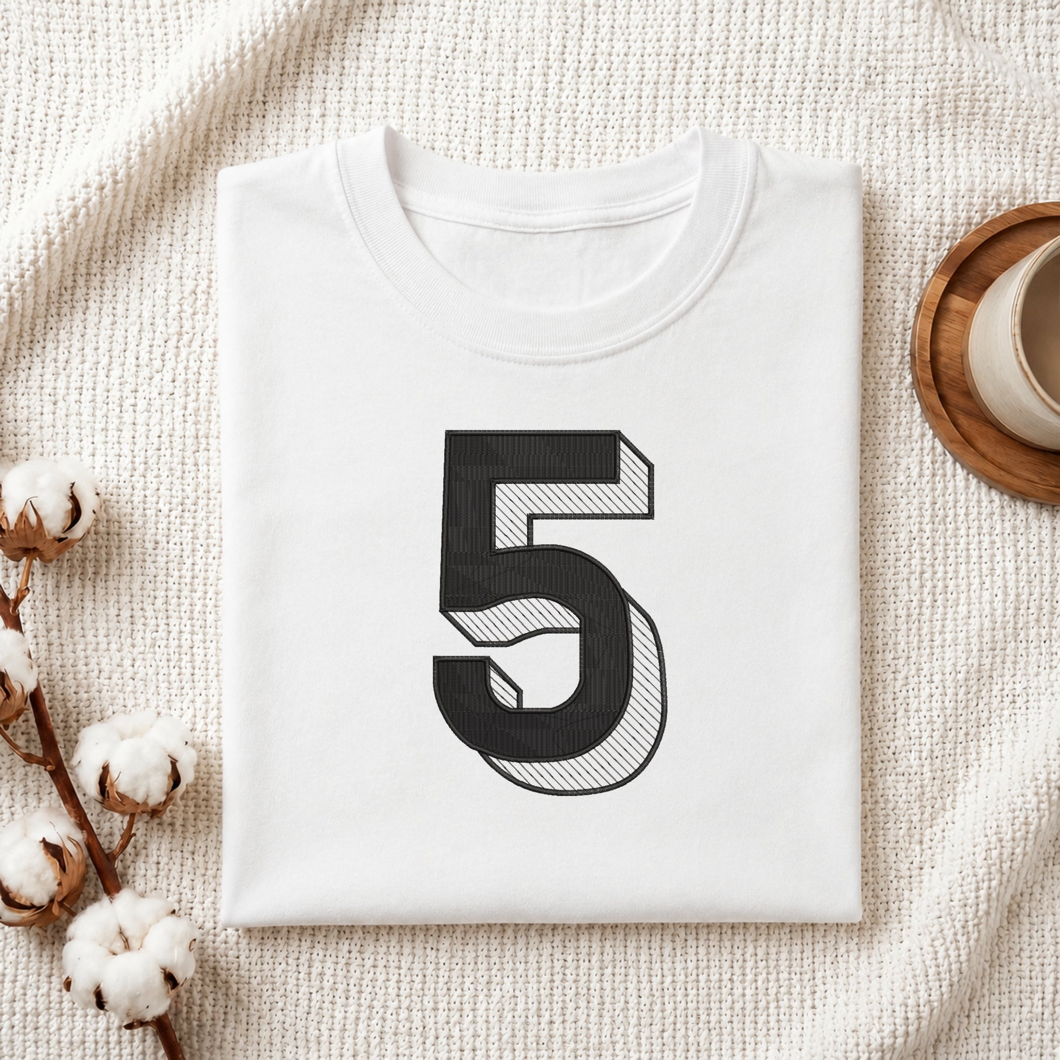A folded white T-shirt with a large black number 5 printed on the front, styled on a textured cream fabric with cotton stems and a small cup in the corners.