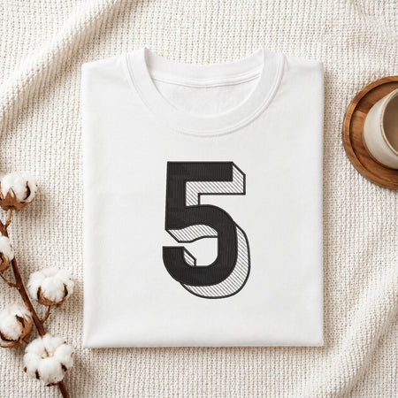A folded white T-shirt with a large black number 5 printed on the front, styled on a textured cream fabric with cotton stems and a small cup in the corners.