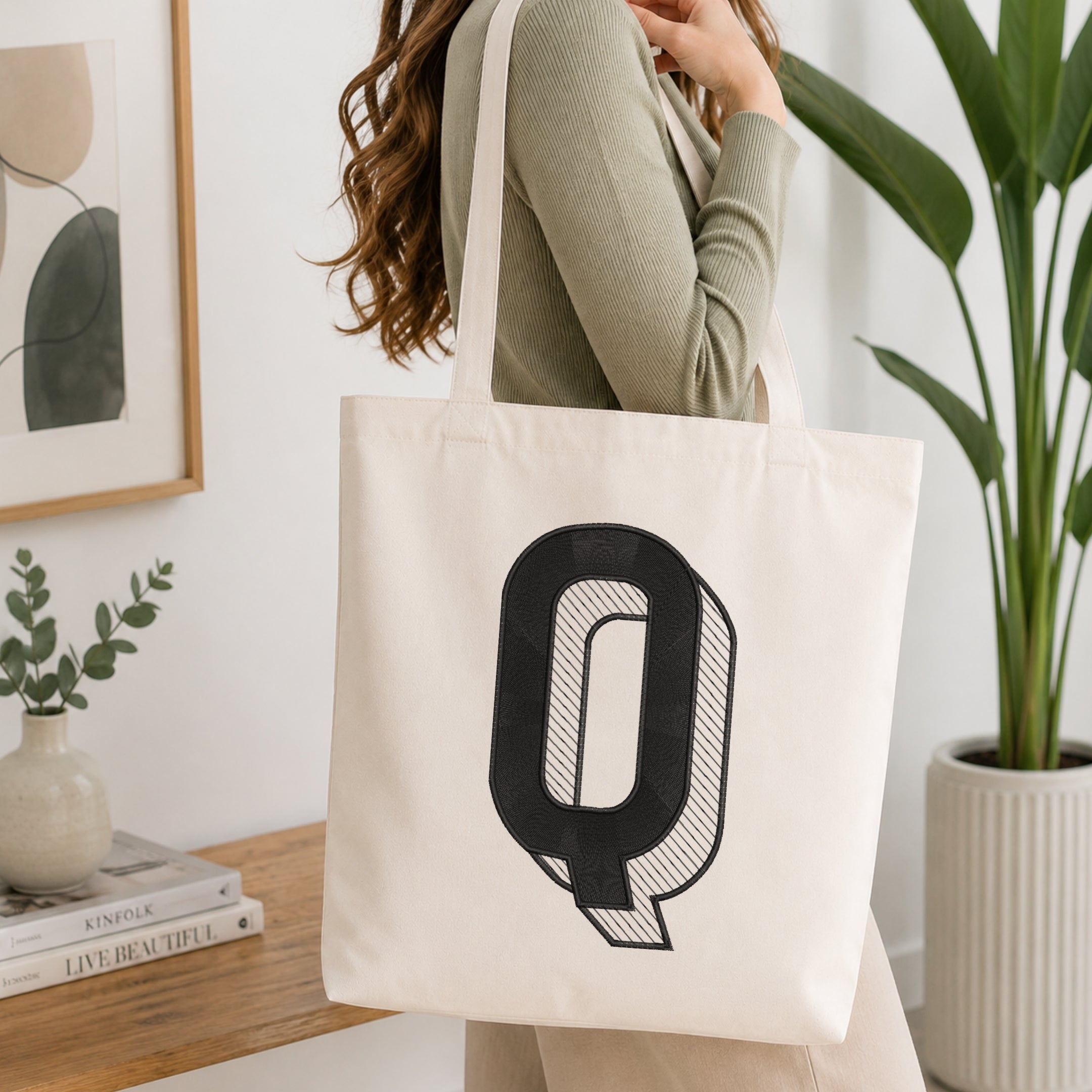 A person holding a natural canvas tote bag printed with an embroidered 3D block letter "Q" design, set against a bright interior room background with green plants.