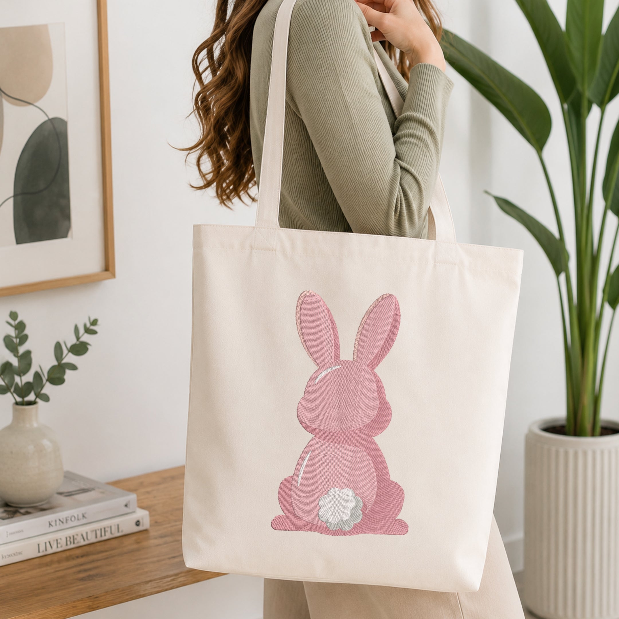 A woman wearing a sage green ribbed sweater carries a cream canvas tote bag with a pink bunny graphic on the front, set in a bright, modern room with a wooden table, framed art, and a potted plant.