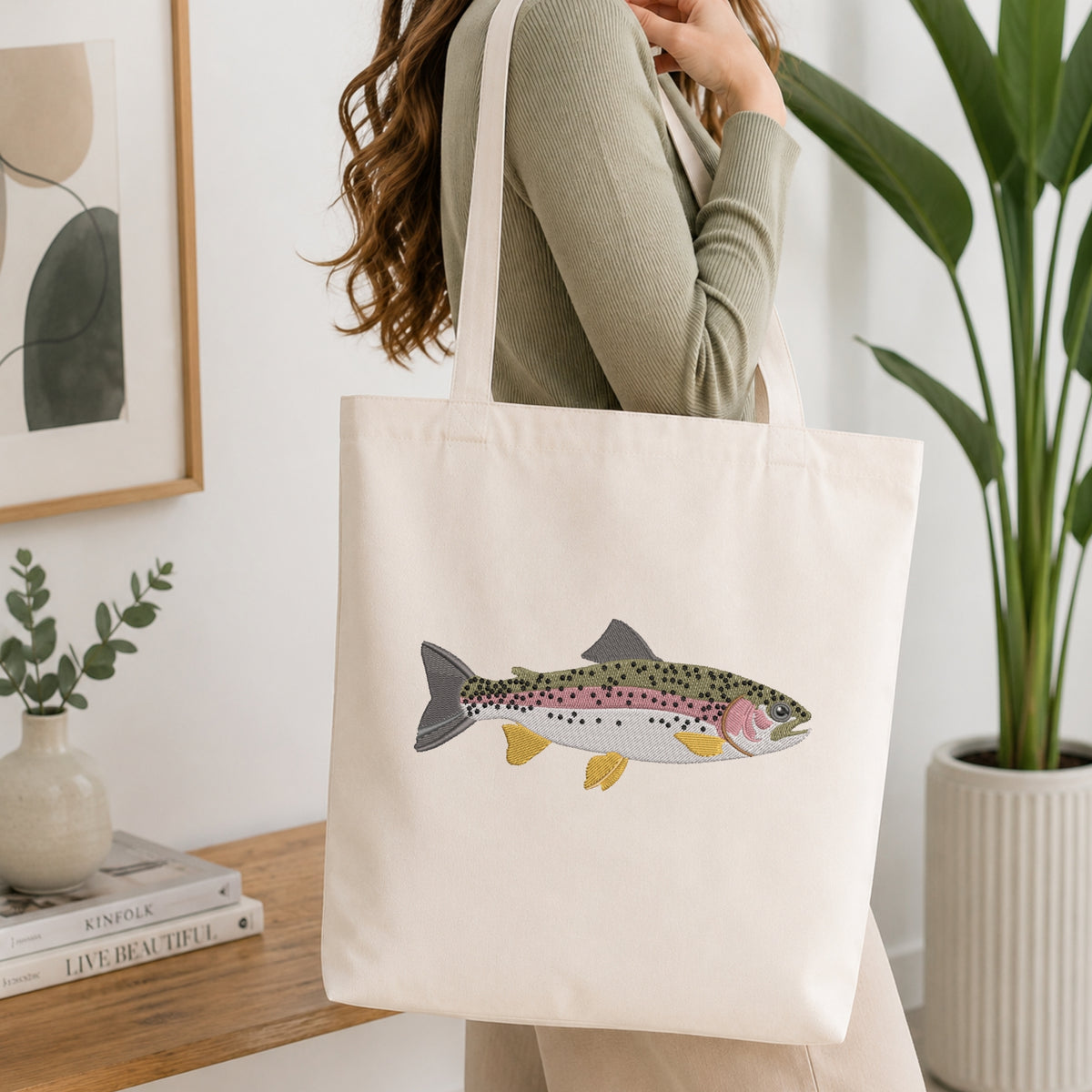 A woman wearing a sage green ribbed sweater carries a cream canvas tote bag with a detailed rainbow trout illustration on the front, set in a bright, modern room with a wooden table, books, and a potted plant.