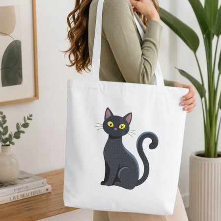 Woman carrying a white natural canvas tote bag featuring an embroidered cute sitting black cat design in a bright, modern interior setting.